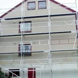 Dwukondygnacyjny dom jednorodzinny w trakcie prac elewacyjnych, otoczony rusztowaniem, z widocznym fragmentem zabezpieczonej folią części budynku.