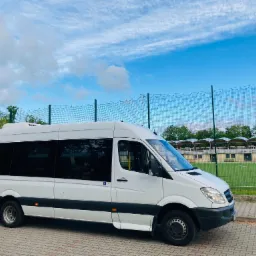 Biały bus Mercedes-Benz Sprinter z przyciemnianymi szybami i symbolem osoby niepełnosprawnej na drzwiach, zaparkowany na kostce brukowej obok boiska sportowego z zieloną murawą i siatką ochronną...
