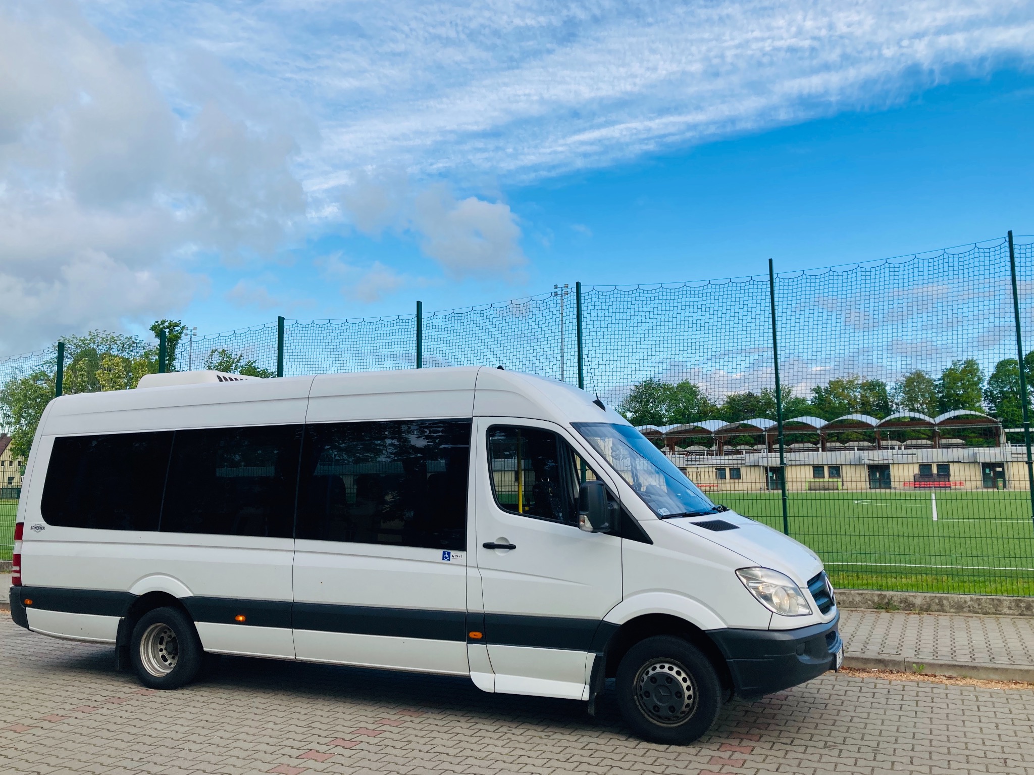 Biały bus Mercedes-Benz Sprinter z przyciemnianymi szybami i symbolem osoby niepełnosprawnej na drzwiach, zaparkowany na kostce brukowej obok boiska sportowego z zieloną murawą i siatką ochronną...