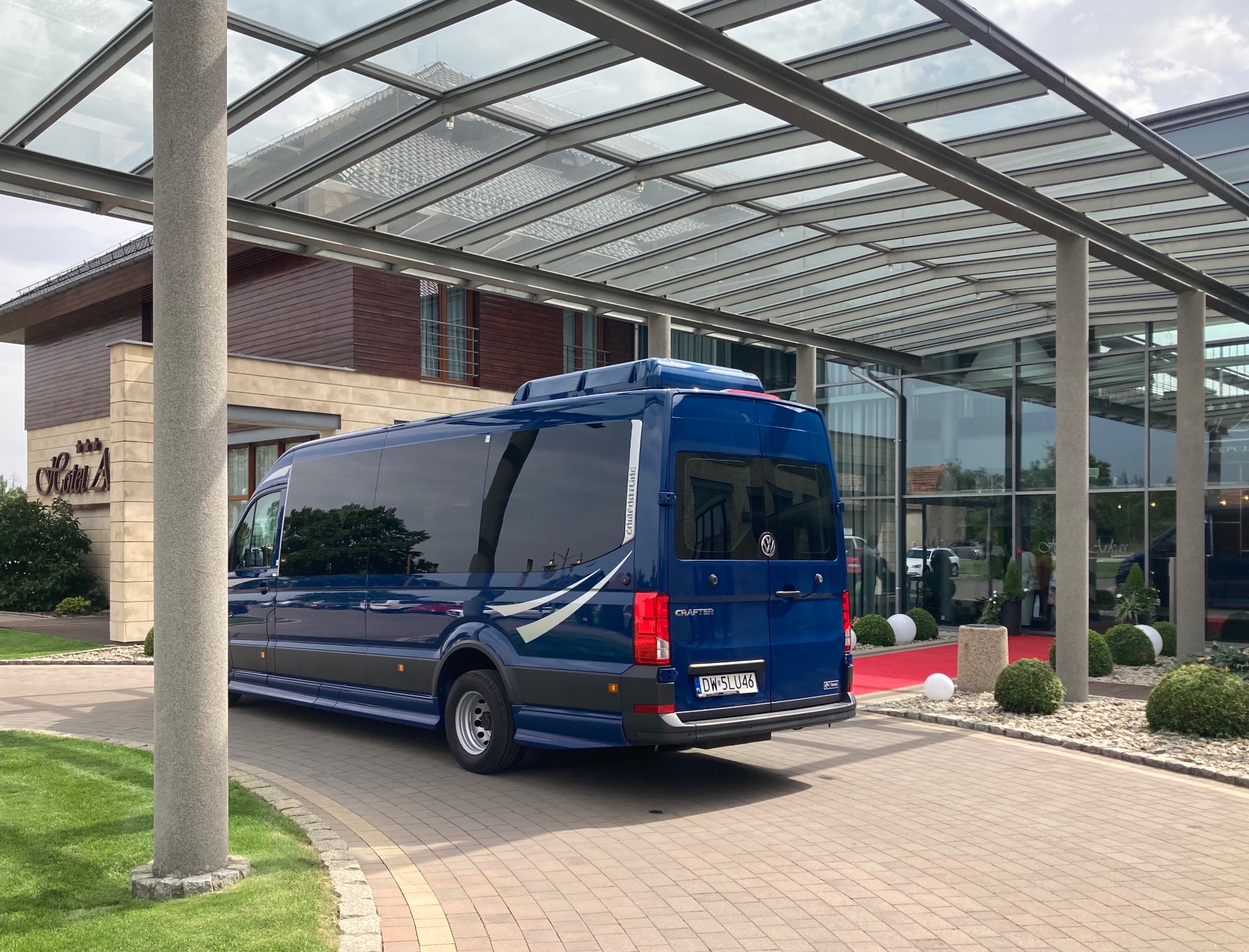 Niebieski bus marki Volkswagen Crafter Comfortline zaparkowany przed wejściem do Hotelu A z czerwonym dywanem, pod przeszklonym zadaszeniem wspartym na kolumnach.