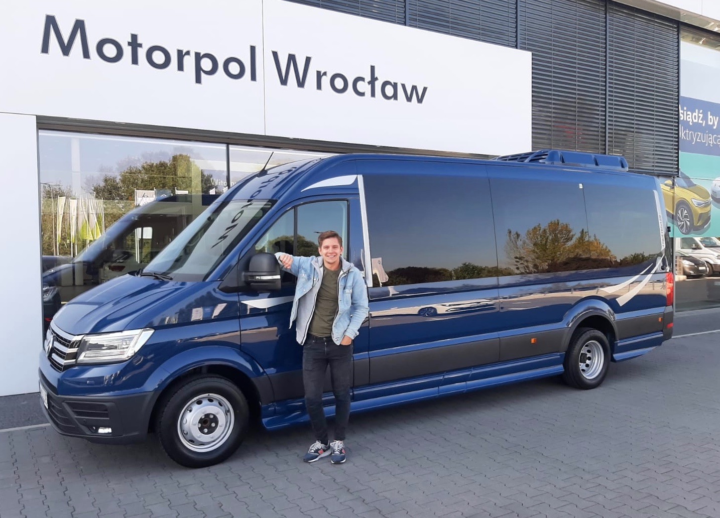 Niebieski bus marki Volkswagen Crafter z wysokim dachem na tle salonu Motorpol Wrocław, obok stoi uśmiechnięty mężczyzna w jeansowej kurtce.