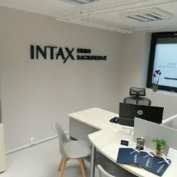 Wnętrze biura rachunkowego z logo firmy 'INTAX' na ścianie, biurka z komputerami, regał z segregatorami i kwiaty w doniczce przy oknie.