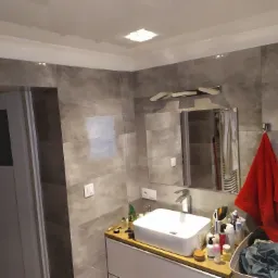 Nowoczesna łazienka z szarymi płytkami imitującymi kamień, białą umywalką nablatową, chromowaną baterią, lustrem z oświetleniem i czerwonym ręcznikiem na wieszaku.