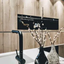 Nowoczesna kuchnia z drewnianymi frontami, czarną armaturą i dekoracyjnymi wazonami z gałązkami bazi na białym blacie.