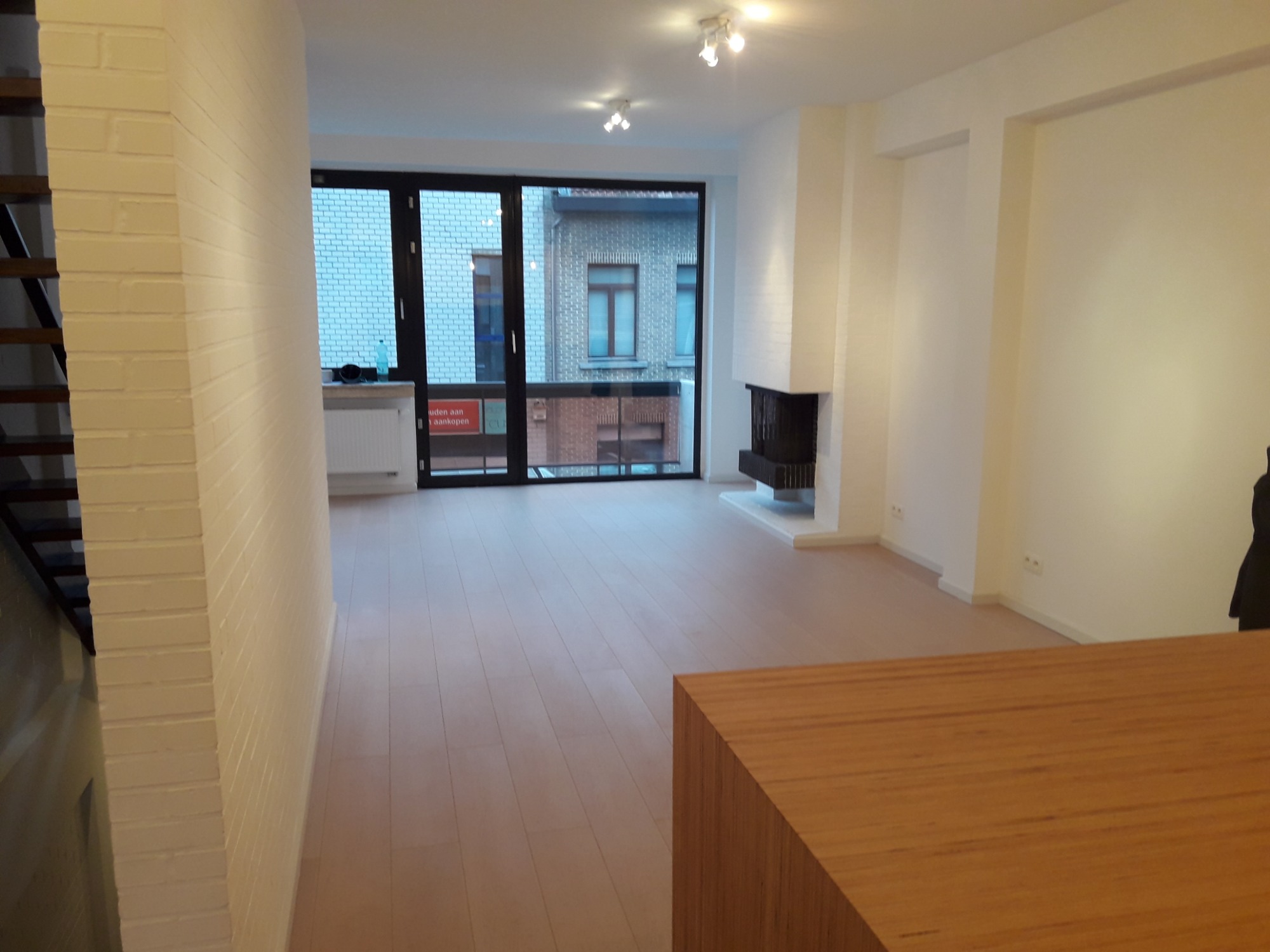 Jasne, przestronne wnętrze z parkietem, białymi ścianami z cegły i kominkiem, widok na balkon przez duże okno.