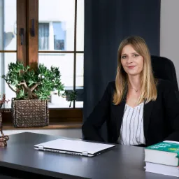 Uśmiechnięta blondynka w czarnej marynarce siedzi za biurkiem w gabinecie. Na biurku posążek Temidy, laptop i stos kodeksów prawnych.