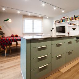 EASY BUILDERS SPÓŁKA Z OGRANICZONĄ ODPOWIEDZIALNOŚCIĄ - Nowoczesna kuchnia z wyspą w odcieniach zieleni, drewniana podłoga, oświetlenie punktowe, stół z czerwoną kratą i choinką w tle, minimalistyczne meble.