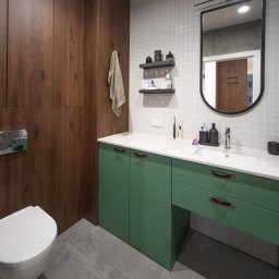 EASY BUILDERS SPÓŁKA Z OGRANICZONĄ ODPOWIEDZIALNOŚCIĄ - Nowoczesna łazienka z drewnianą zabudową, zieloną szafką pod umywalką z uchwytami ze skóry, białą umywalką, lustrem w czarnej ramie i płytkami na ścianach.