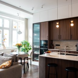 EASY BUILDERS. Projekt z Wykończeniem - Nowoczesne, otwarte wnętrze z salonem, jadalnią i kuchnią. Drewniana podłoga w jodełkę, szara sofa, okrągły stół z beżowymi krzesłami, ciemne szafki kuchenne i marmurowy blat z wyspą.