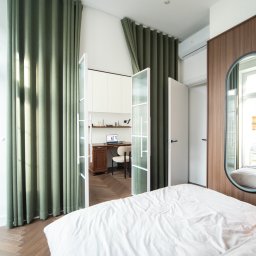 EASY BUILDERS. Projekt z Wykończeniem - Nowoczesna sypialnia z białym łóżkiem, zielonymi zasłonami i drewnianą podłogą w jodełkę. Za szklanymi drzwiami wydzielony kącik do pracy z biurkiem i laptopem. Owalne lustro.