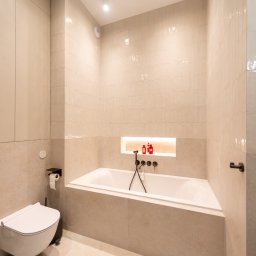 EASY BUILDERS. Projekt z Wykończeniem - Nowoczesna łazienka z beżowymi płytkami o dwóch fakturach, wanną w zabudowie, podświetlaną wnęką z czerwonymi akcesoriami i czarnymi, matowymi armaturami.
