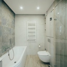 EASY BUILDERS. Projekt z Wykończeniem - Nowoczesna łazienka z wanną, podwieszanym WC i szklaną kabiną prysznicową. Szare płytki ścienne, jedna z dekoracyjnym wzorem. Podłoga imitująca drewno, okrągłe lustro i biały grzejnik.