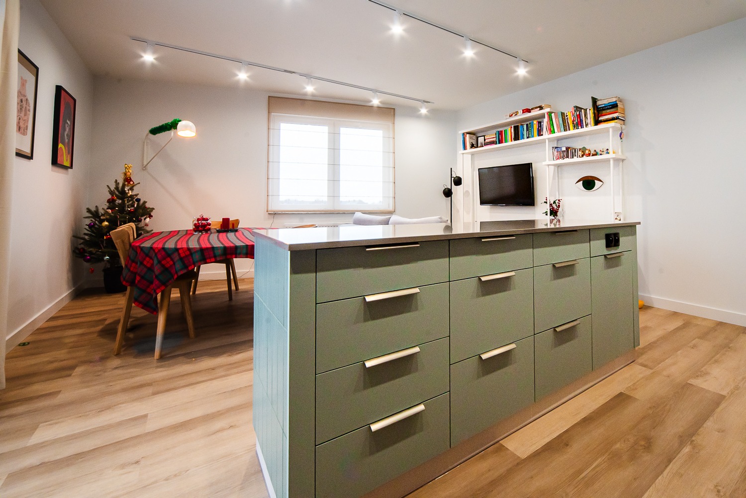 Nowoczesna kuchnia z wyspą w odcieniach zieleni, drewniana podłoga, oświetlenie punktowe, stół z czerwoną kratą i choinką w tle, minimalistyczne meble.