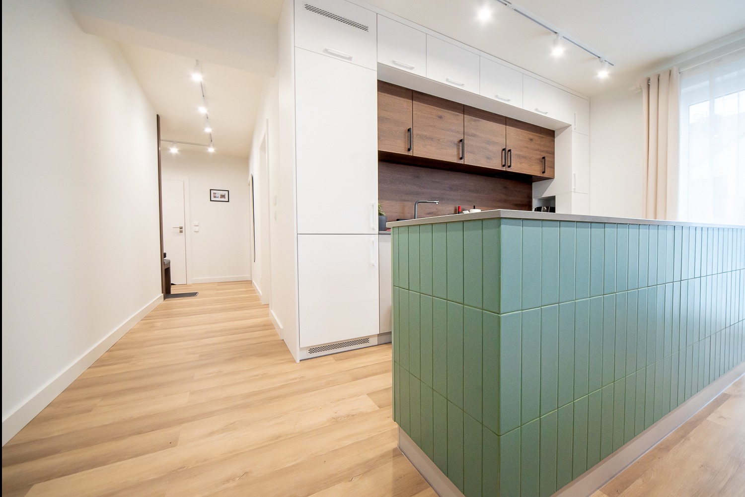 Nowoczesna kuchnia z wyspą wykończoną pionowymi, zielonymi płytkami, drewniane szafki wiszące i białe szafki stojące, jasna podłoga i oświetlenie punktowe.