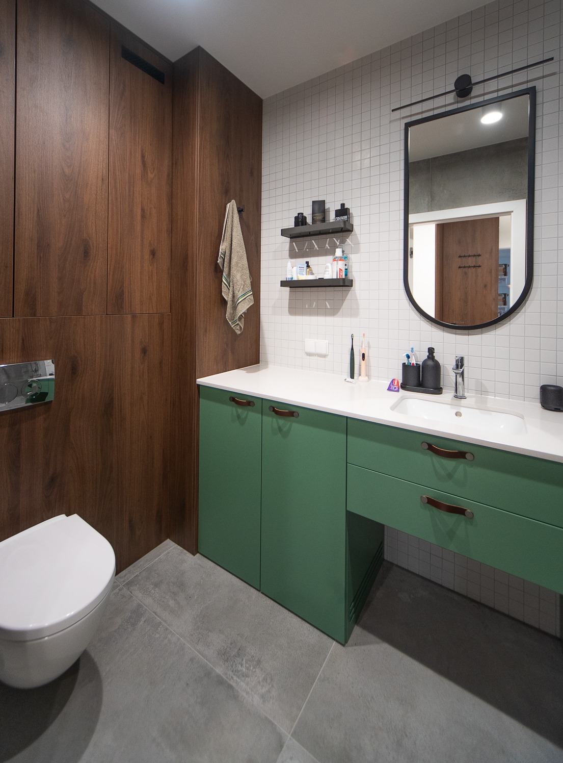 Nowoczesna łazienka z drewnianą zabudową, zieloną szafką pod umywalką z uchwytami ze skóry, białą umywalką, lustrem w czarnej ramie i płytkami na ścianach.