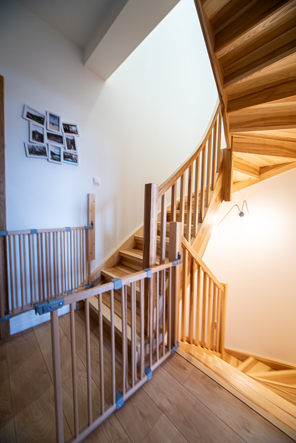 Drewniane schody kręcone z barierką zabezpieczającą dla dzieci, na ścianie galeria zdjęć, jasne wnętrze.