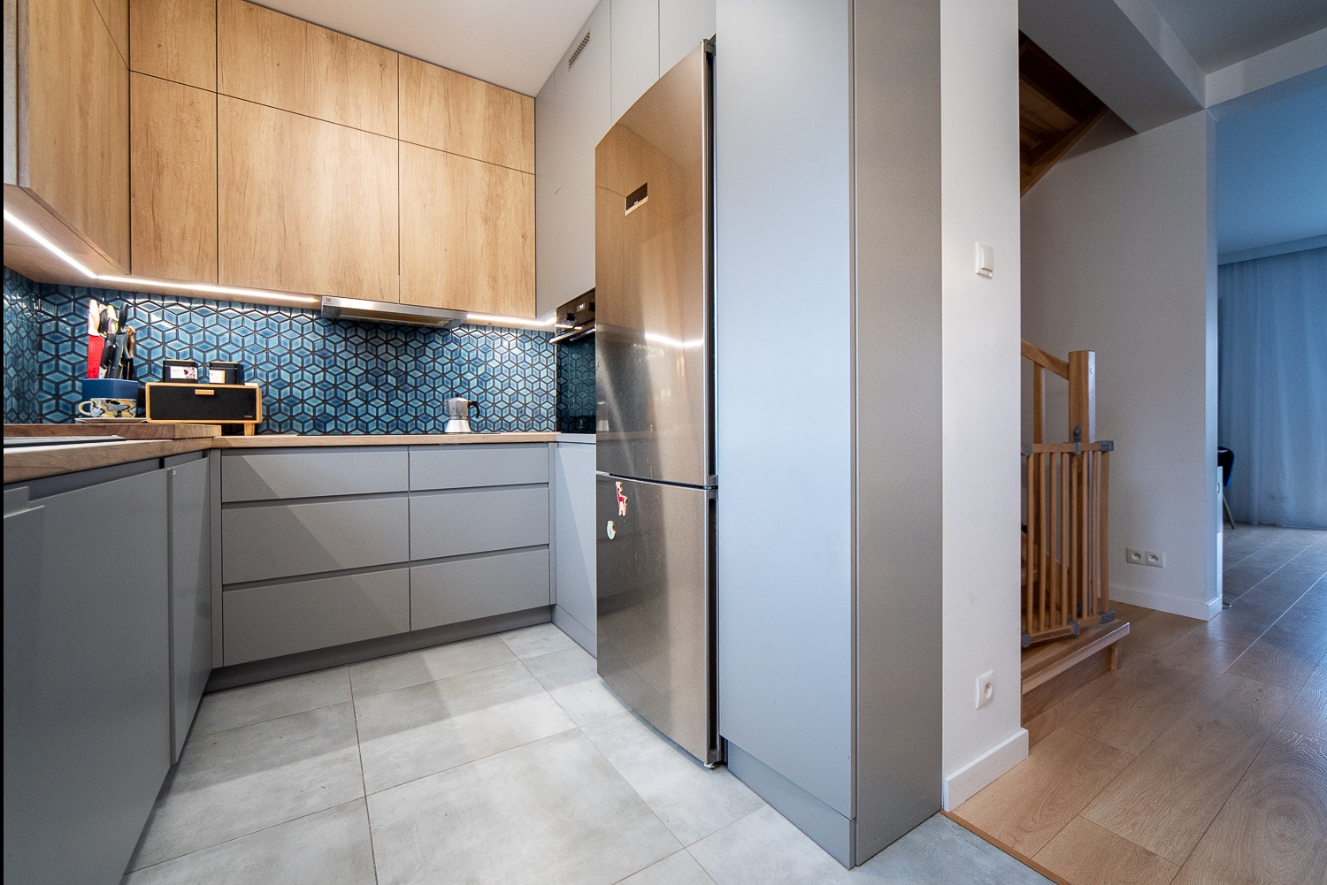 Nowoczesna kuchnia z drewnianymi szafkami górnymi, szarymi szafkami dolnymi i geometrycznymi, niebieskimi płytkami na ścianie. Widoczna lodówka ze stali nierdzewnej i fragment schodów z drewnianą...