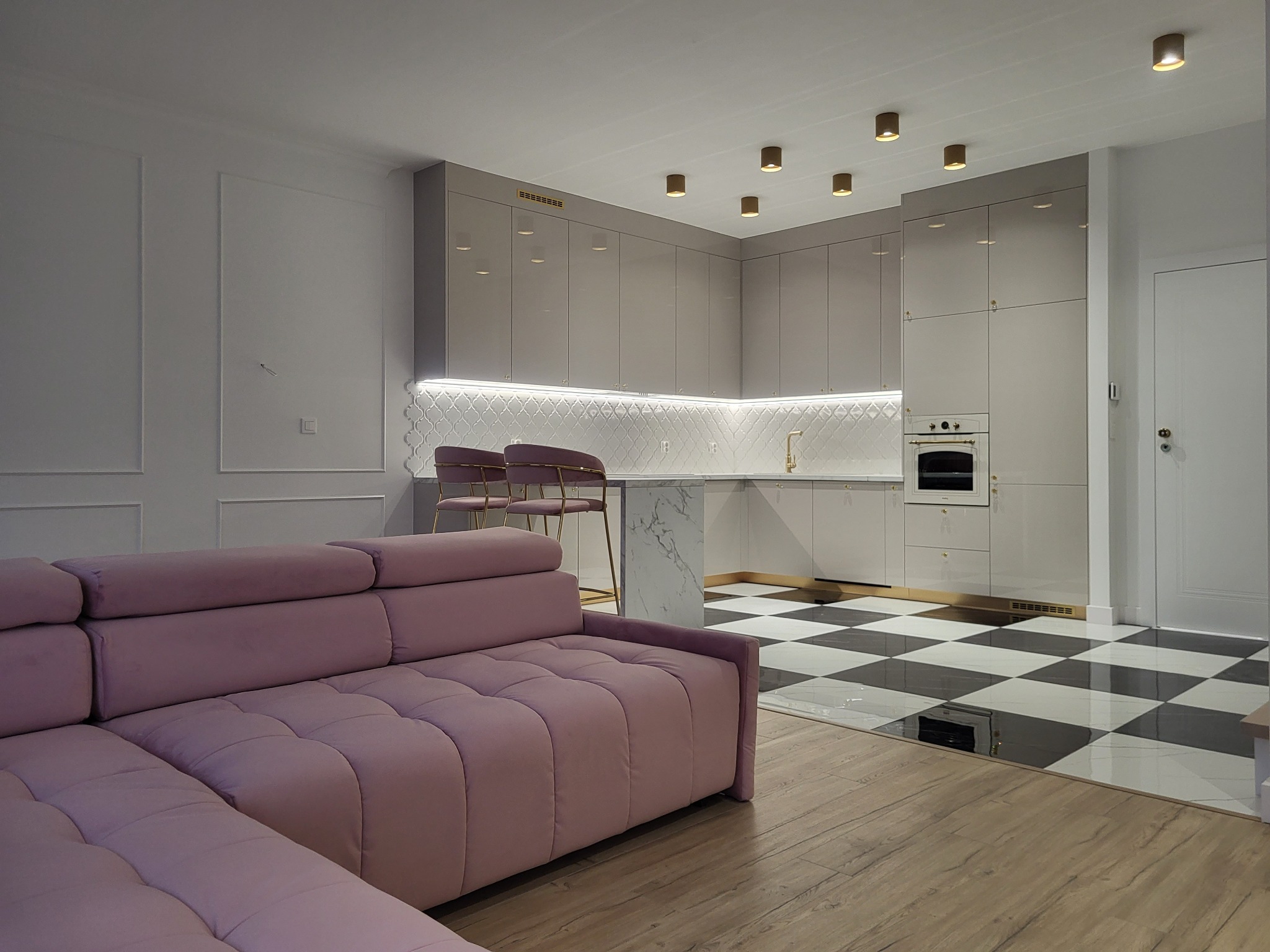 Nowoczesne wnętrze: szara kuchnia z marmurowym blatem i złotymi akcentami, fioletowa sofa w salonie, czarno-biała podłoga w szachownicę, drewniana podłoga w salonie.