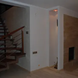 Nowoczesne wnętrze z drewnianymi schodami z metalową balustradą, ścianą pokrytą dekoracyjną fakturą i obudowanym kominkiem z jasnego kamienia.