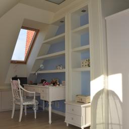 Firma Budowlana Styl-Bud - Pokój na poddaszu z białymi meblami: biurko z krzesłem, regał z półkami i szafa. Widoczne okno dachowe i zabawki na biurku.