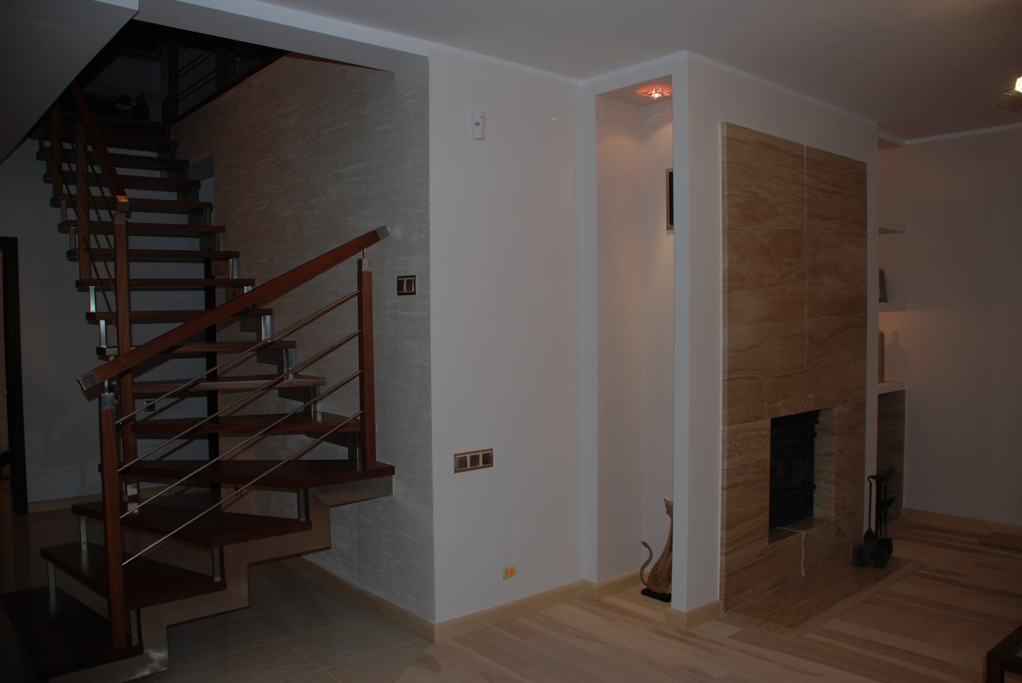 Nowoczesne wnętrze z drewnianymi schodami z metalową balustradą, ścianą pokrytą dekoracyjną fakturą i obudowanym kominkiem z jasnego kamienia.