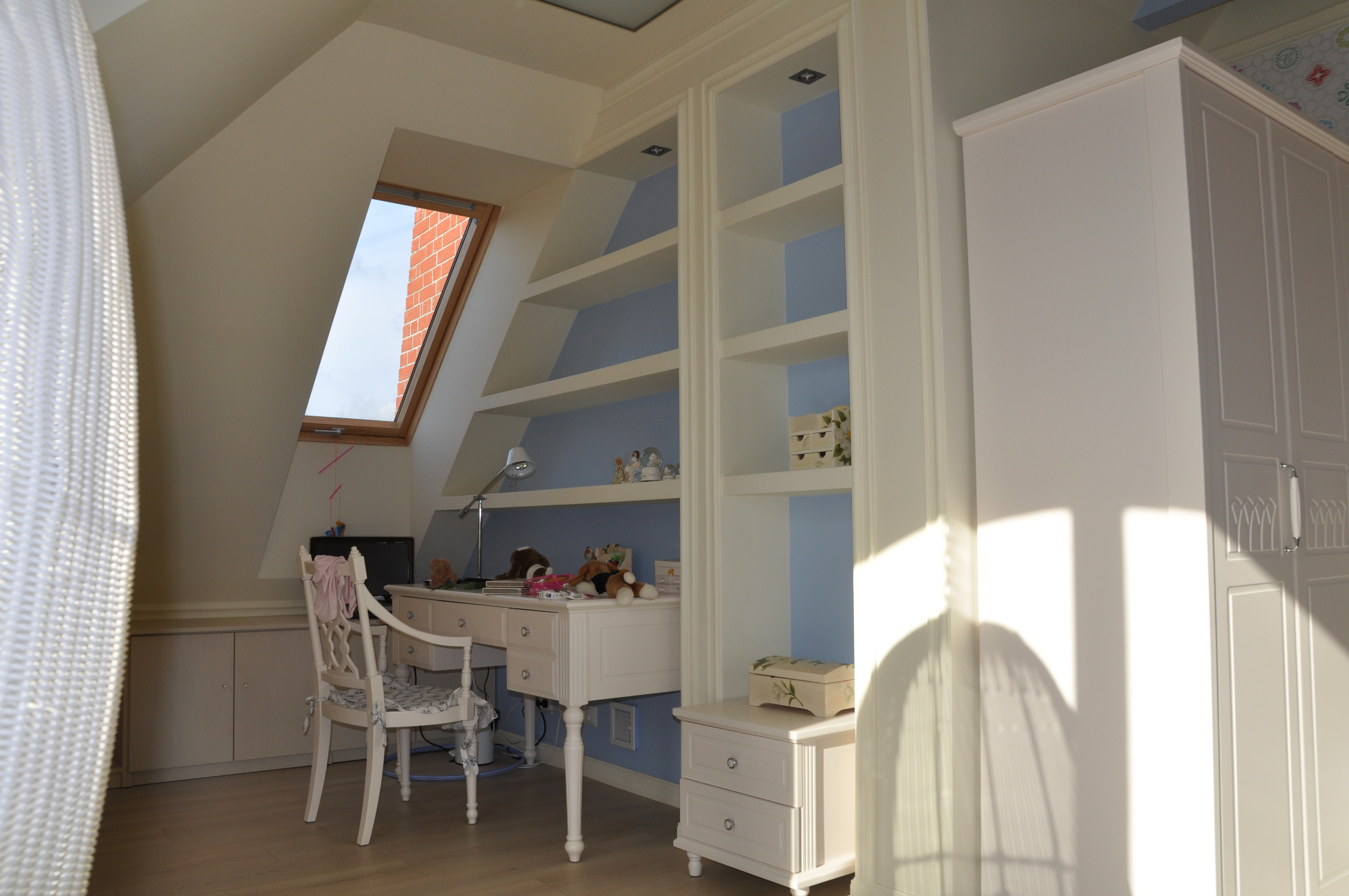 Pokój na poddaszu z białymi meblami: biurko z krzesłem, regał z półkami i szafa. Widoczne okno dachowe i zabawki na biurku.