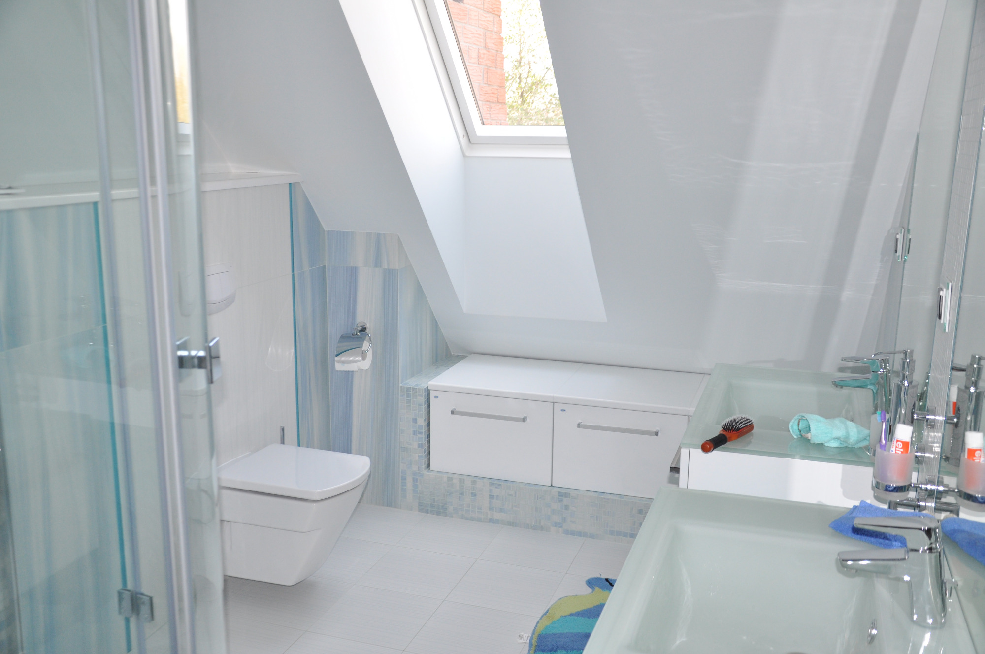 Jasna łazienka na poddaszu z oknem dachowym, białą ceramiką, umywalkami z przezroczystego szkła i niebieskimi akcentami na płytkach i tekstyliach.