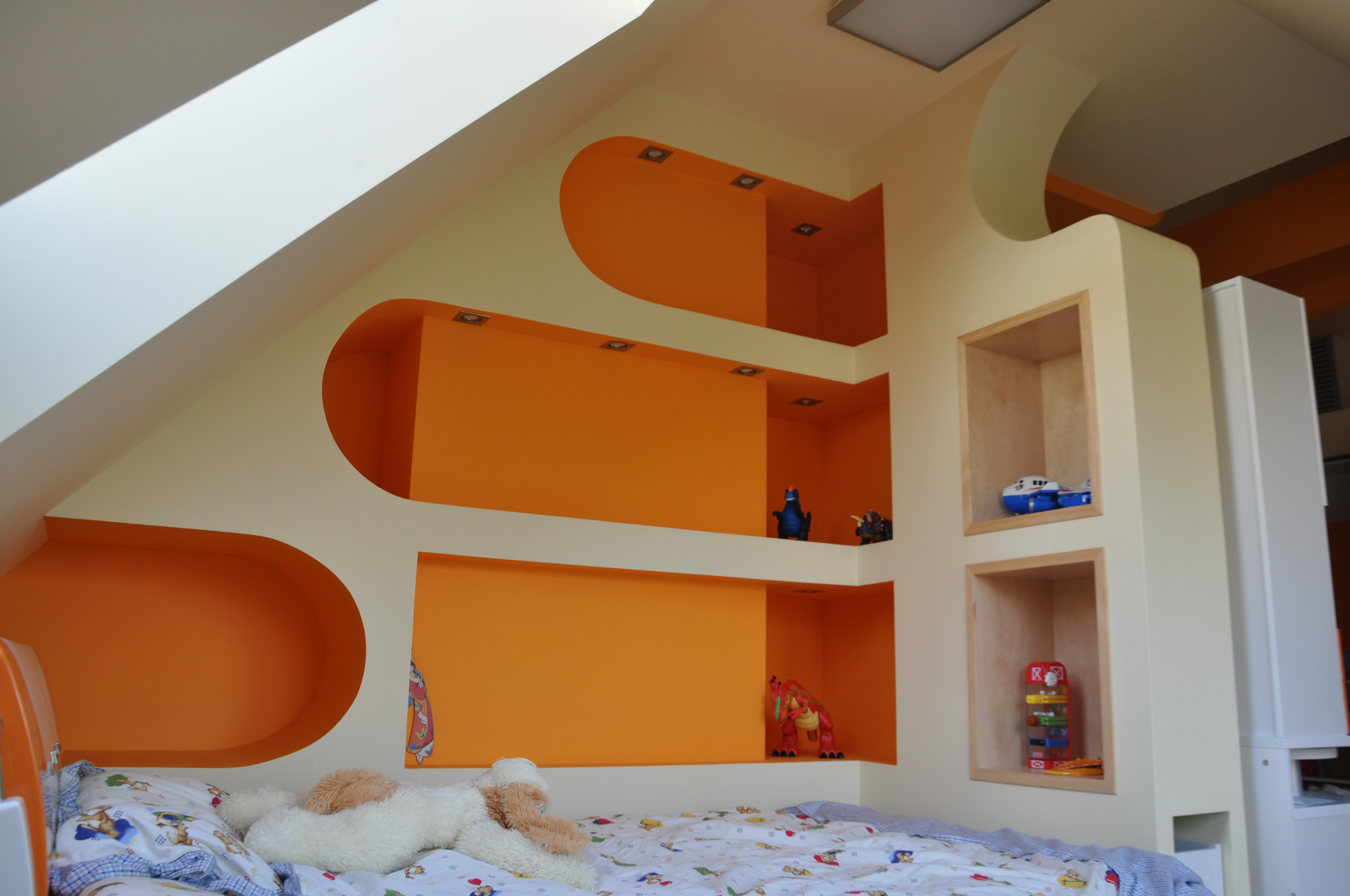 Pokój dziecięcy na poddaszu z pomarańczowymi wnękami w ścianie, oświetleniem punktowym i półkami z zabawkami, widok na łóżko z pościelą w dziecięce wzory.