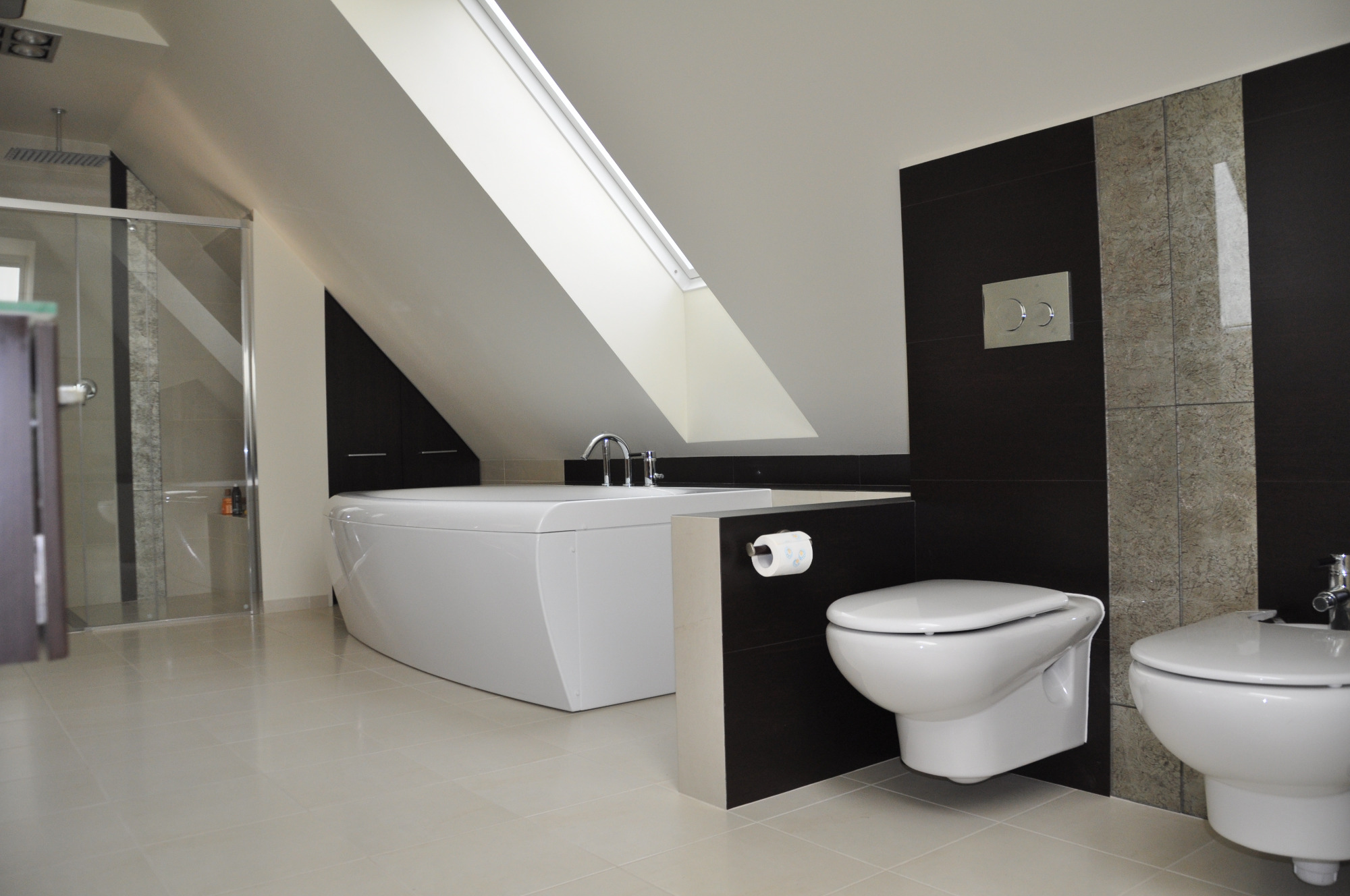 Nowoczesna łazienka na poddaszu z białą, prostokątną wanną, kabiną prysznicową ze szklanymi drzwiami i wiszącą toaletą z czarnymi panelami. Okno dachowe wpuszcza naturalne światło.