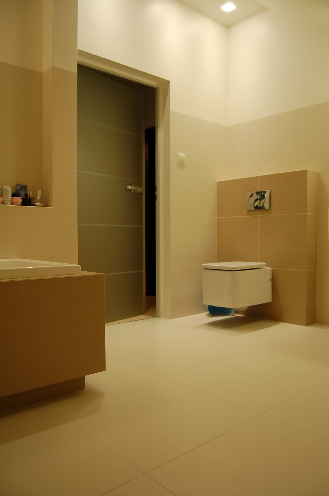 Nowoczesna łazienka z podwieszaną toaletą, beżowymi płytkami i szklanymi drzwiami, oświetlona ciepłym światłem.