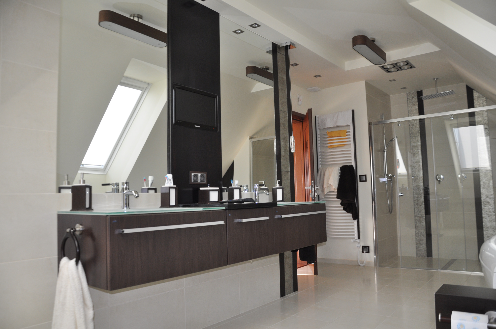 Nowoczesna łazienka na poddaszu z oknem dachowym, umywalką na szafce z ciemnego drewna, lustrem z wbudowanym telewizorem i kabiną prysznicową ze szklanymi drzwiami.