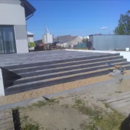 Schody z kostki brukowej prowadzące do budynku, widoczny zagęszczarka wibracyjna i piasek, w tle zabudowania gospodarcze.