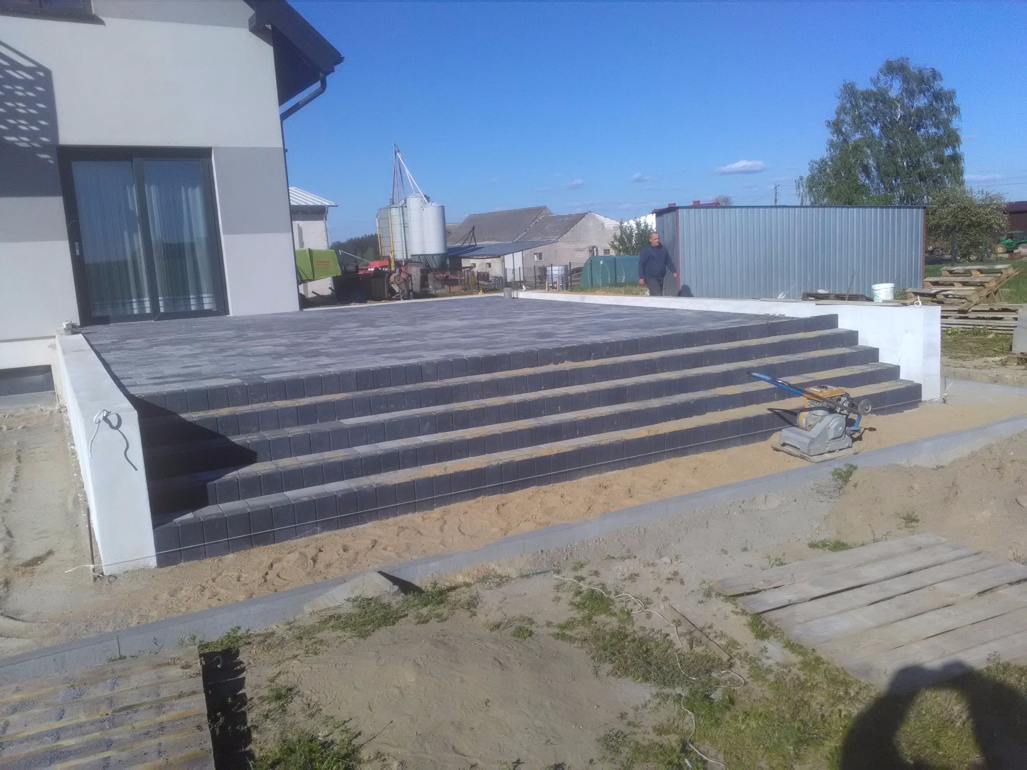 Schody z kostki brukowej prowadzące do budynku, widoczny zagęszczarka wibracyjna i piasek, w tle zabudowania gospodarcze.