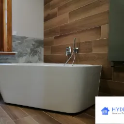 Nowoczesna, wolnostojąca wanna z chromowaną baterią, na tle ściany wyłożonej brązowymi płytkami imitującymi drewno i szarej marmurowej ściany, z widocznym logo firmy Hydrokris w prawym dolnym rogu.
