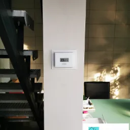 Biały termostat Vaillant zamontowany na ścianie, obok metalowych schodów kratowych. W tle widoczne refleksy słoneczne na ścianie i biurko z dokumentami.