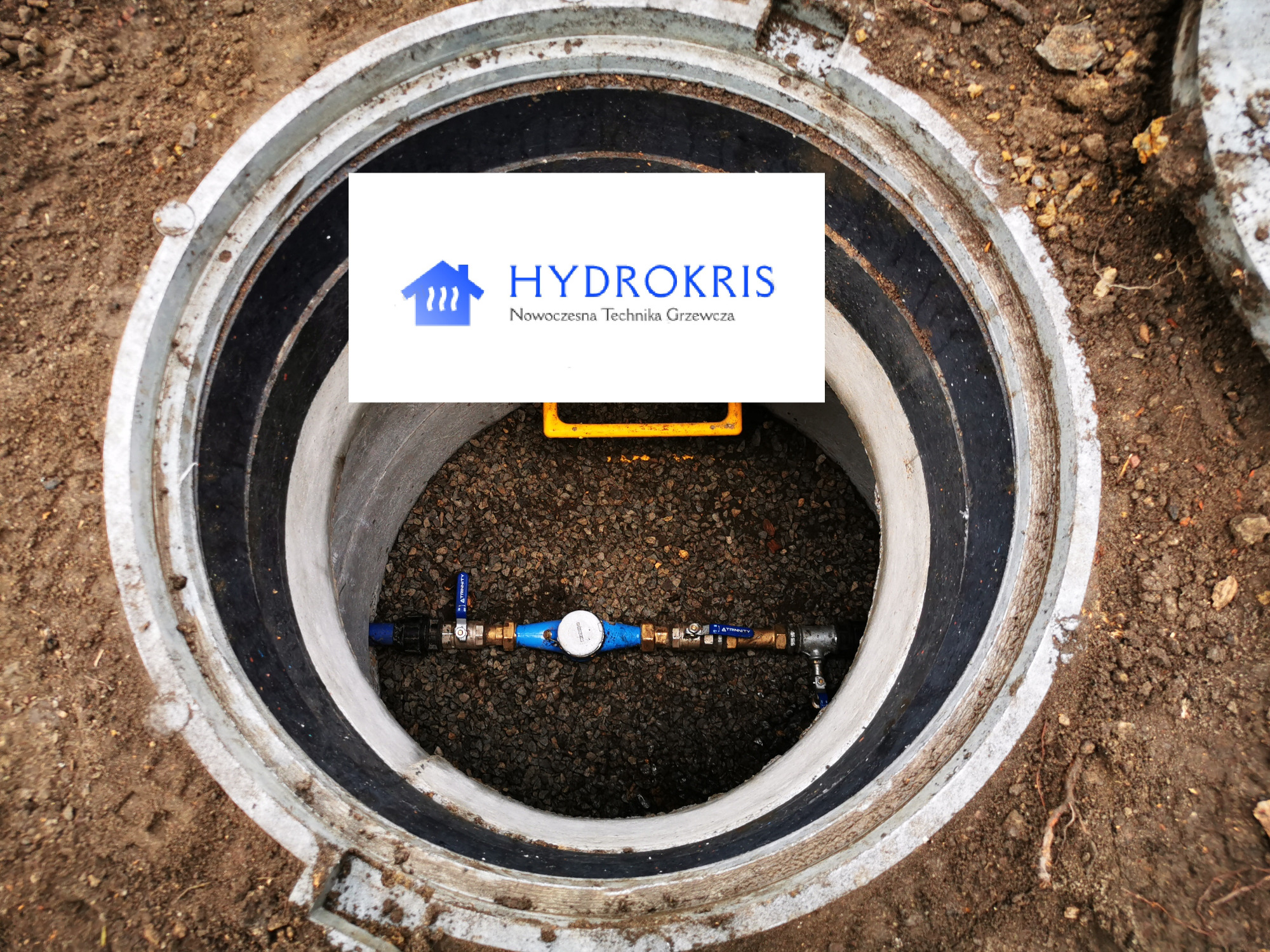 Studzienka rewizyjna z licznikiem wody, widoczny fragment instalacji wodnej z zaworami i logo firmy 'Hydrokris' na białym tle.