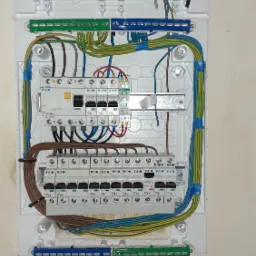 Wnętrze białej skrzynki rozdzielczej z zainstalowanymi bezpiecznikami EATON, przewodami różnej grubości i kolorów, oraz listwami zaciskowymi, zabezpieczonymi niebieską taśmą izolacyjną.