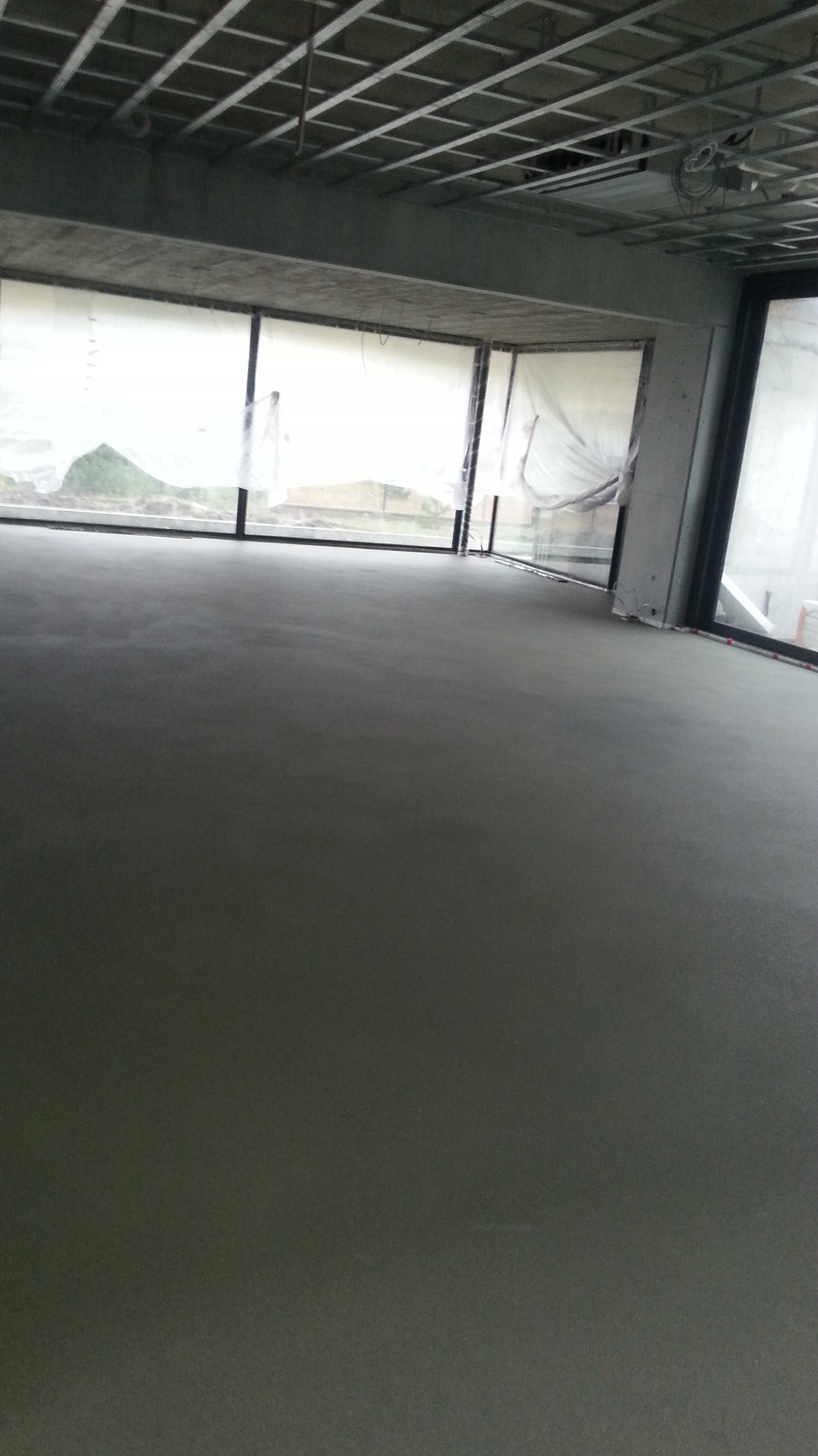 Wylana, gładka posadzka jastrychowa w surowym wnętrzu z dużymi oknami, zabezpieczonymi folią. Widoczna metalowa konstrukcja sufitu podwieszanego.