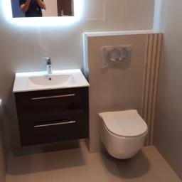 Prace wykończeniowe - Dominik Turczyn - Nowoczesna łazienka w szarościach z podwieszaną toaletą, umywalką z szafką w kolorze ciemnego brązu i lustrem z podświetleniem LED, w którym odbija się osoba robiąca zdjęcie.