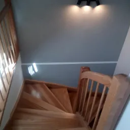 Drewniane schody z balustradą, oświetlone trzema reflektorami na szarej ścianie, widok z góry.