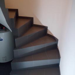 Prace wykończeniowe - Dominik Turczyn - Spiralne schody wyłożone szarymi płytkami ceramicznymi, widoczne z dołu, z bojlerem po lewej stronie.
