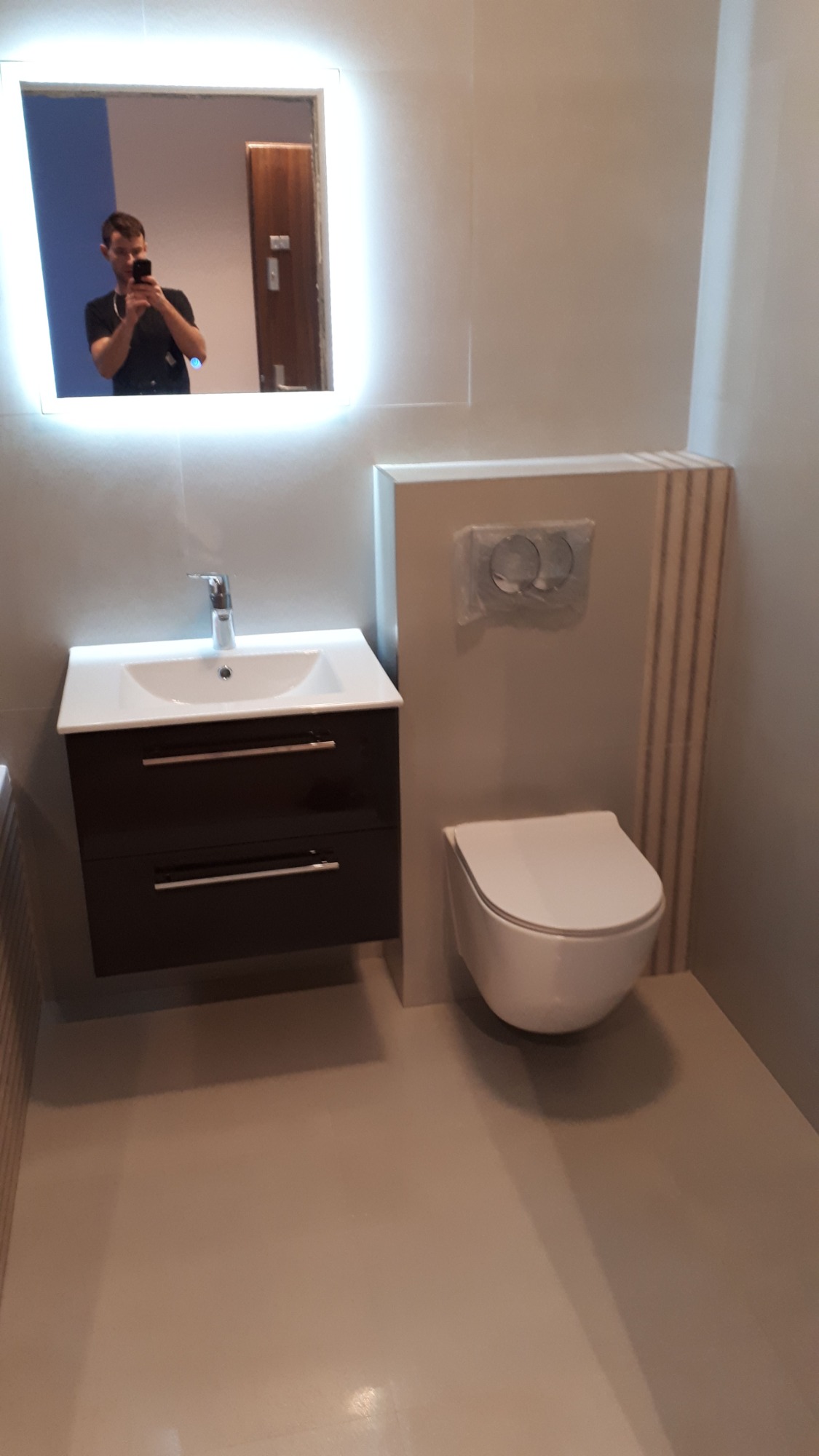 Nowoczesna łazienka w szarościach z podwieszaną toaletą, umywalką z szafką w kolorze ciemnego brązu i lustrem z podświetleniem LED, w którym odbija się osoba robiąca zdjęcie.