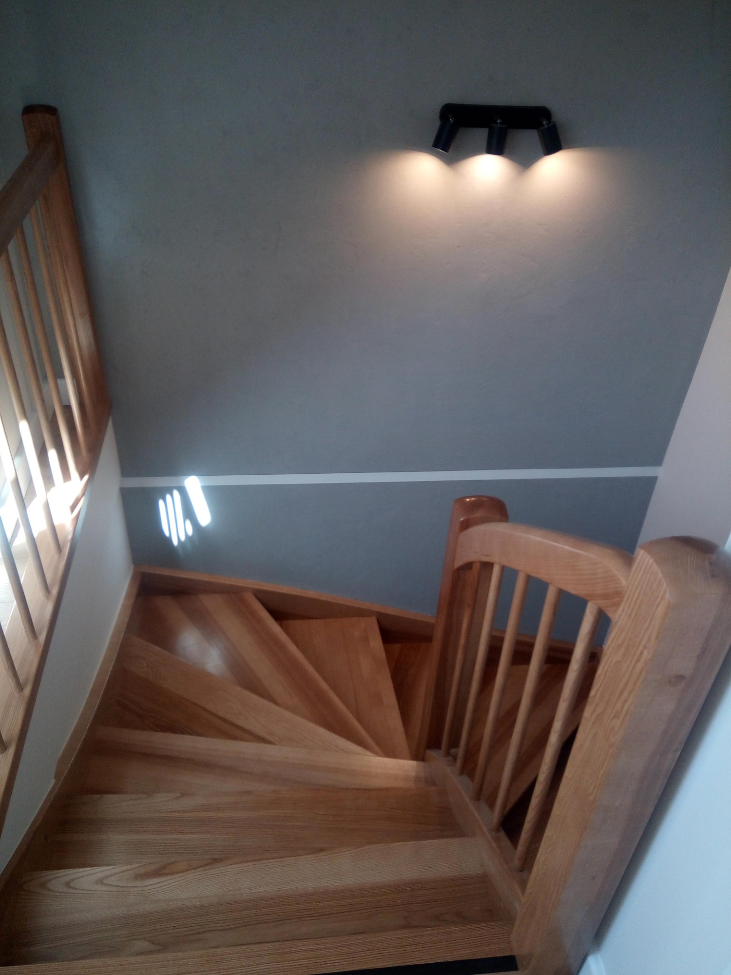 Drewniane schody z balustradą, oświetlone trzema reflektorami na szarej ścianie, widok z góry.