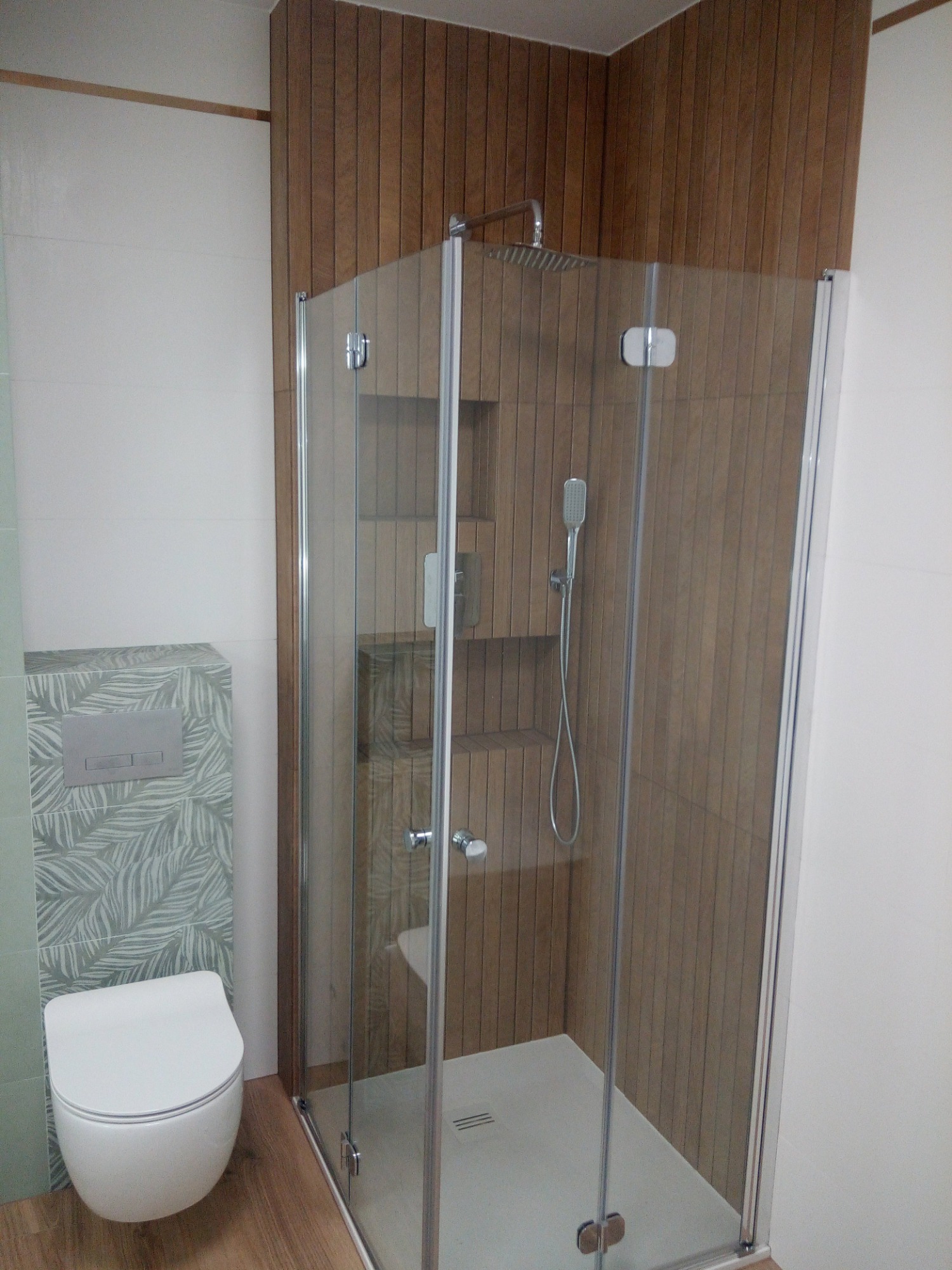 Nowoczesna kabina prysznicowa z deszczownicą i półkami w obudowie, wisząca toaleta z wzorem liści, drewnopodobne płytki na ścianie i podłodze.