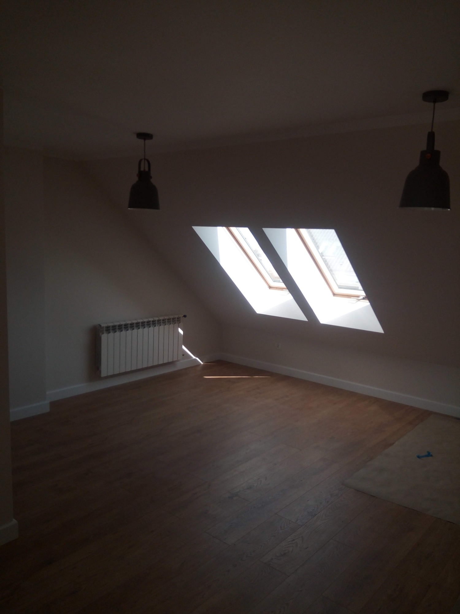 Poddasze z dwoma oknami dachowymi, drewnianą podłogą i grzejnikiem pod oknem, oświetlone przez dwie czarne lampy wiszące.