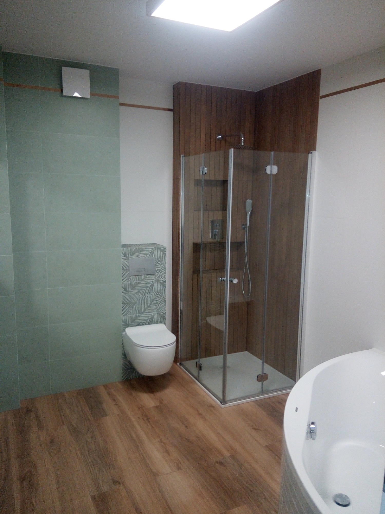 Nowoczesna łazienka z kabiną prysznicową, podwieszaną toaletą z wzorzystą ścianką i drewnopodobną podłogą, oświetlona światłem z górnego okna.