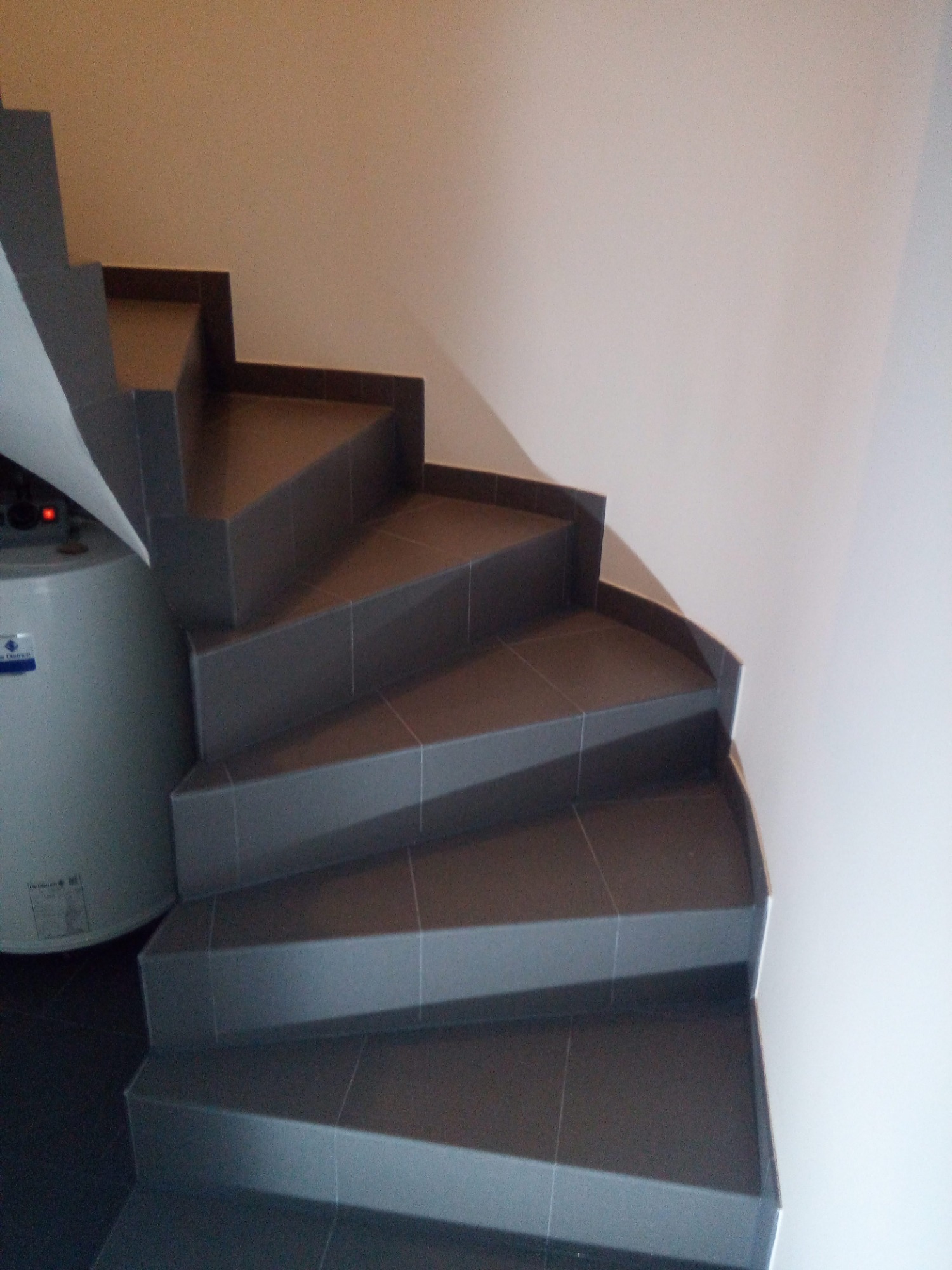 Spiralne schody wyłożone szarymi płytkami ceramicznymi, widoczne z dołu, z bojlerem po lewej stronie.