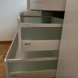 Wysuwane szuflady z białymi frontami i szklanymi panelami, widoczne logo Blum na każdym froncie, zamontowane w szafie z białymi drzwiami, częściowo otwarte, ukazujące wnętrze.