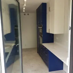 Nowoczesna kuchnia z granatowymi szafkami dolnymi i białymi górnymi, marmurowym blatem i oświetleniem punktowym, widziana z perspektywy wejścia.