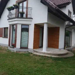 Dwukondygnacyjny dom z białą elewacją, brązowymi akcentami okiennymi i drewnopodobną okładziną, balkon z kwiatami i schody wejściowe z resztkami zaprawy.